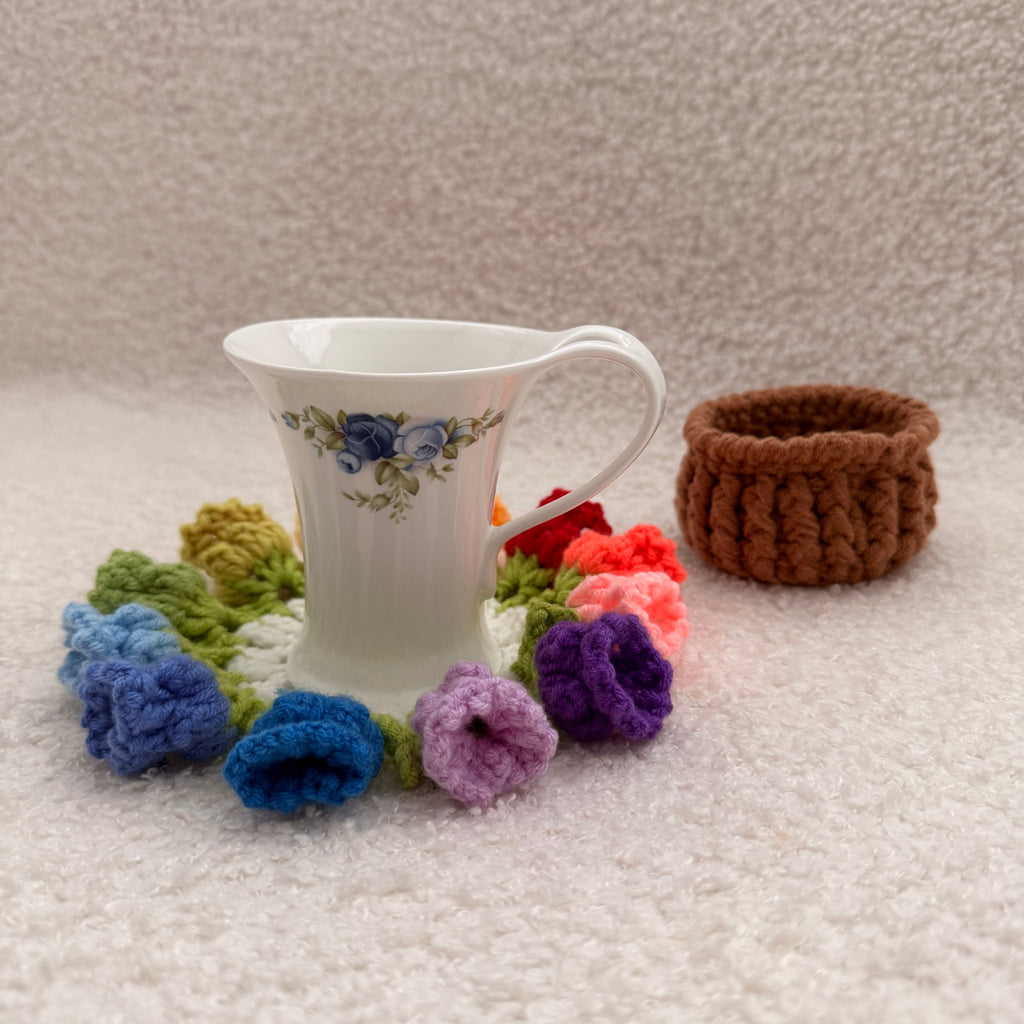 White teacup with floral design surrounded by colorful knitted flowers on a textured surface.