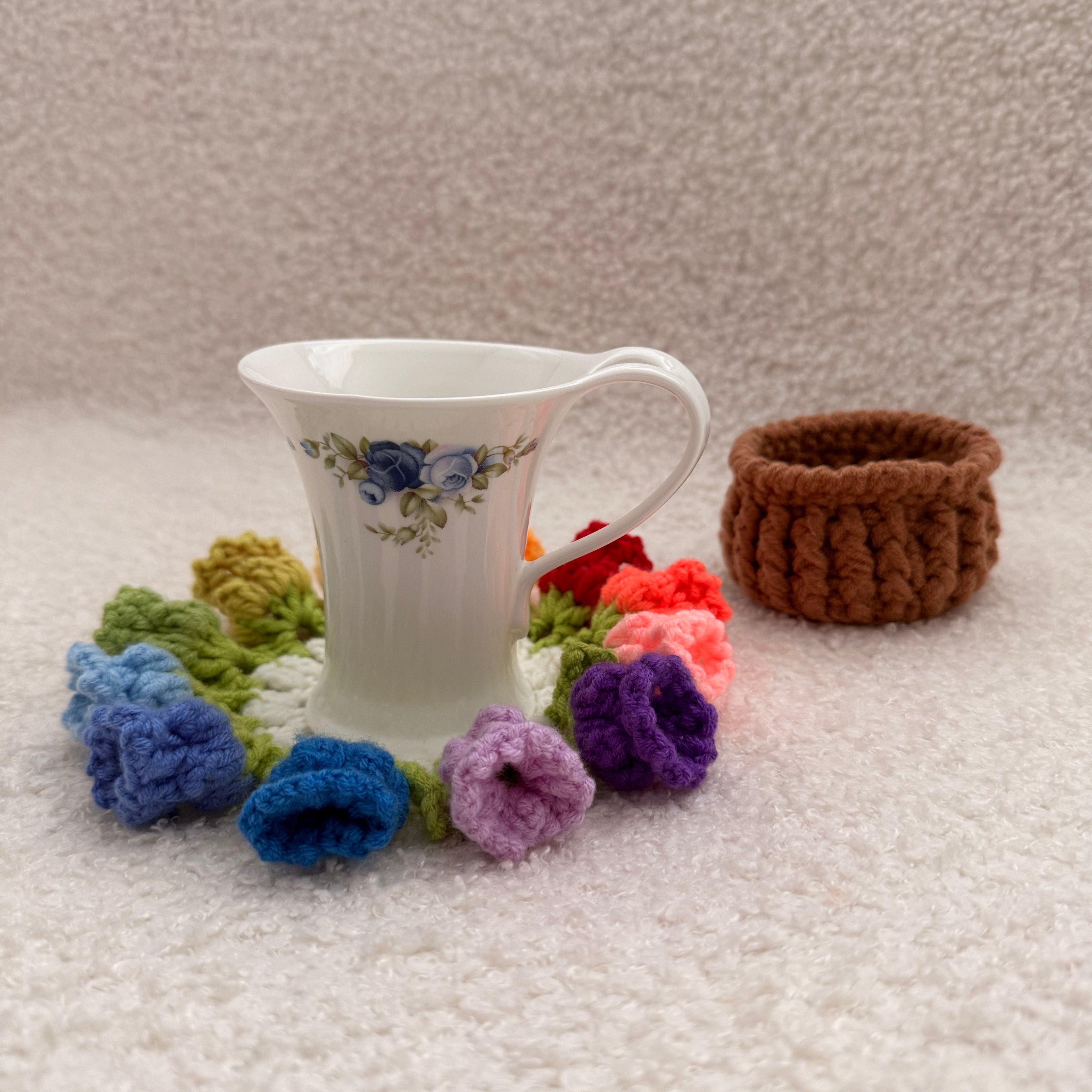 White teacup with floral design surrounded by colorful knitted flowers on a textured surface.